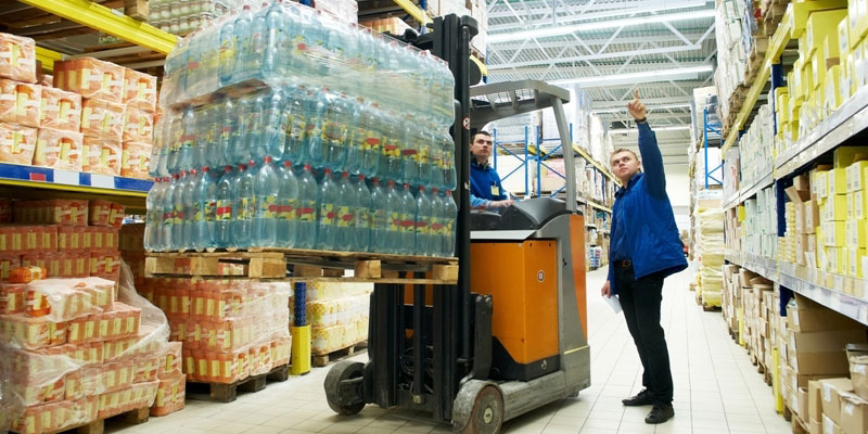 Insurance for warehouses Two warehouse workers discussing storage options for the water bottles being carried by a forklift