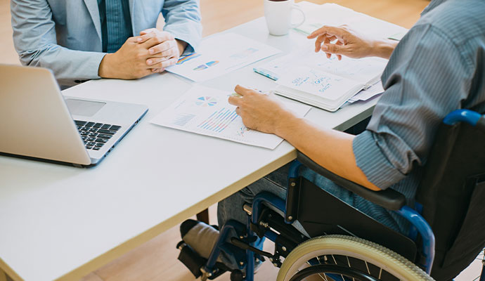 Employee using wheelchair collaborating with coworker on charts and laptop at desk
